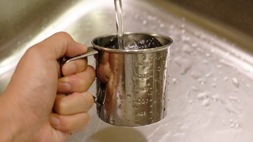 Hand holding a stainless steel measuring jug under a running kitchen tap to measure water flow rate in litres per minute.
