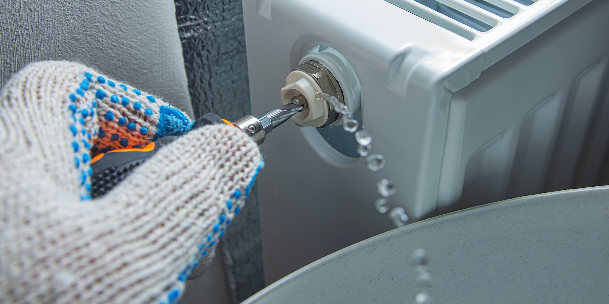 Gloved hand using a radiator bleed key to bleed a panel radiator as water drains into a bucket.